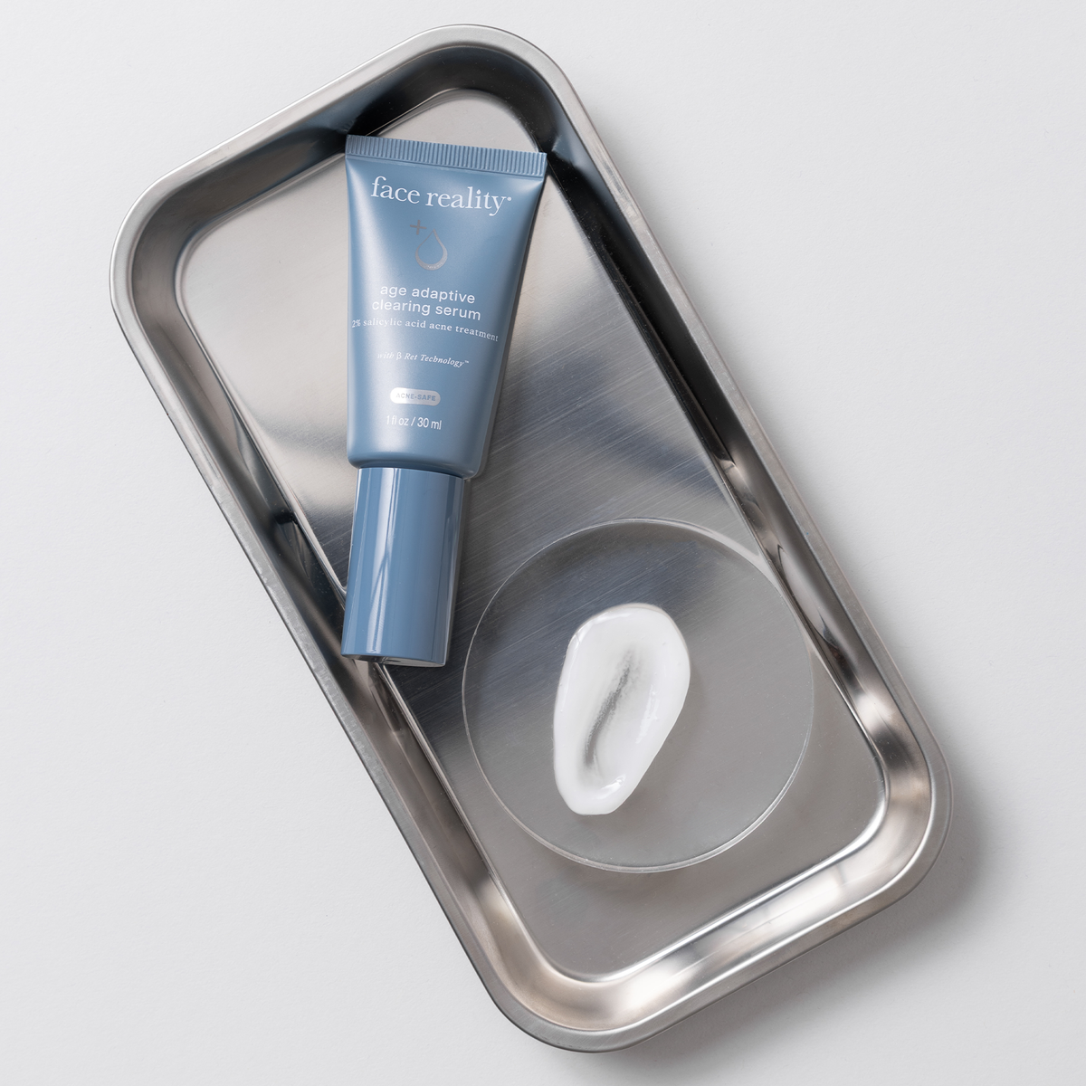 Face Reality Age Adaptive Clearing Serum tube on a metal tray beside a drop of the serum